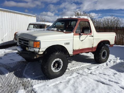 1984 Ford Bronco - A white and red Ford Bronco is parked in the snow on a sunny day. The vehicle has large off-road tires and appears to be modified.
