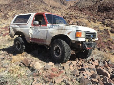 1984 Ford Bronco - A weathered white and red Ford Bronco with off-road tires is parked on a rocky, dry hillside.