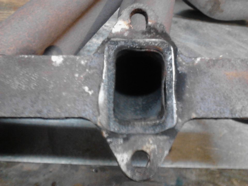 Close-up of a rusty exhaust manifold flange with a square opening and bolt holes.