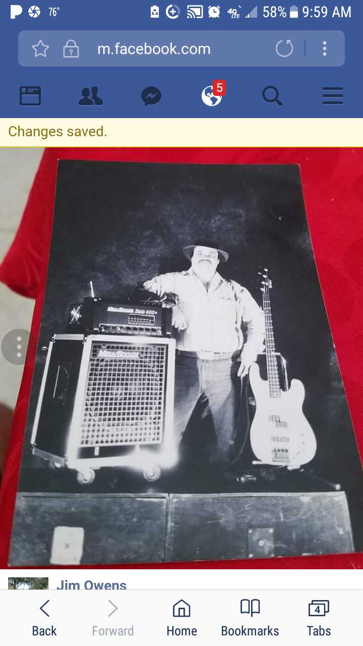 Phone screenshot of a black-and-white photo of a man posing with bass amps and a guitar