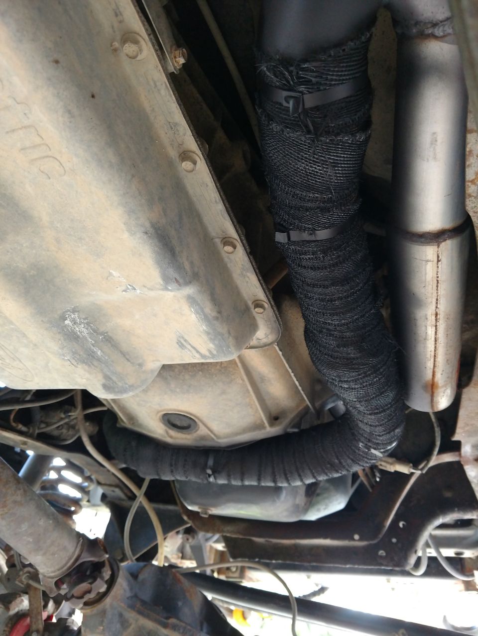 Underside view of vehicle showing exhaust pipes wrapped in black heat tape and a transmission pan