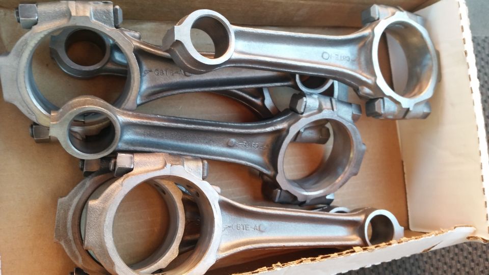 Four metal engine connecting rods in a cardboard box. Some rods have part numbers stamped on them