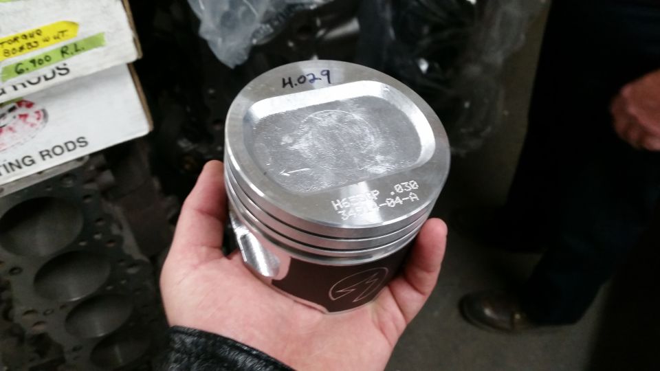 Hand holding a piston with markings '4.029' and 'H655TP .030 3400-04-A'. Box of connecting rods in background