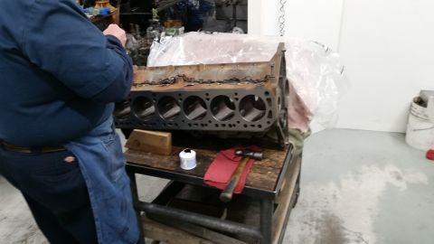 1981 Ford Bronco - A person in a blue shirt and denim apron works on a V8 engine block on a workbench. The engine block has six cylinders visible and appears…