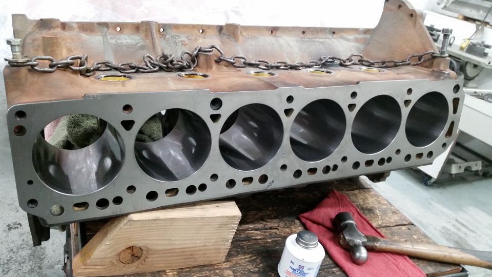 V8 engine block with six cylinders, secured by a chain, resting on wood.