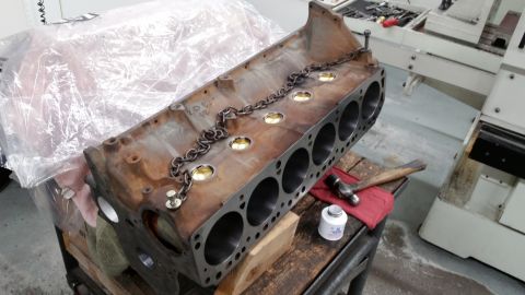 1981 Ford Bronco - A rusty V8 engine block is shown on a workbench, covered partially by plastic sheeting. A metal chain is draped across the top of the block…