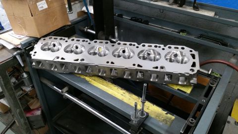 1981 Ford Bronco - A clean, polished aluminum engine cylinder head with six combustion chambers is positioned on a workbench with various tools and machinery…