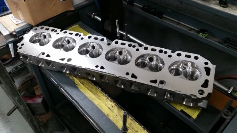 1981 Ford Bronco - A clean, silver aluminum engine cylinder head with six combustion chambers is shown lying on a workbench. The cylinder head has intake and…