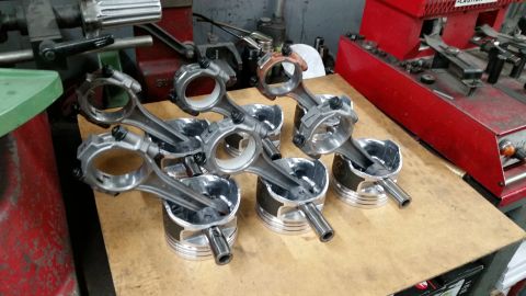1981 Ford Bronco - Several sets of connecting rods are attached to pistons, resting on a wooden surface in what appears to be a workshop. A red machine is vis…