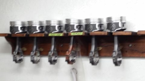 1981 Ford Bronco - A set of eight pistons with connecting rods are displayed on a wooden rack mounted on a white wall. Each piston has a dark finish with a ci…