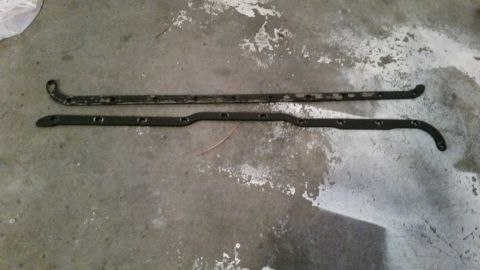 1981 Ford Bronco - Two dark-colored, L-shaped metal brackets with multiple holes are laid out on a textured concrete floor. The brackets appear to be automoti…