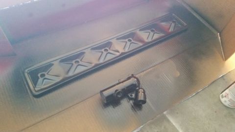 1981 Ford Bronco - A black valve cover and two other small parts are spray painted inside a cardboard box. The valve cover has a raised, star-like pattern wit…