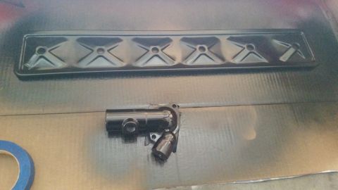 1981 Ford Bronco - A black painted valve cover and a black painted thermostat housing are laid out on a cardboard surface. A roll of blue painter's tape is vi…