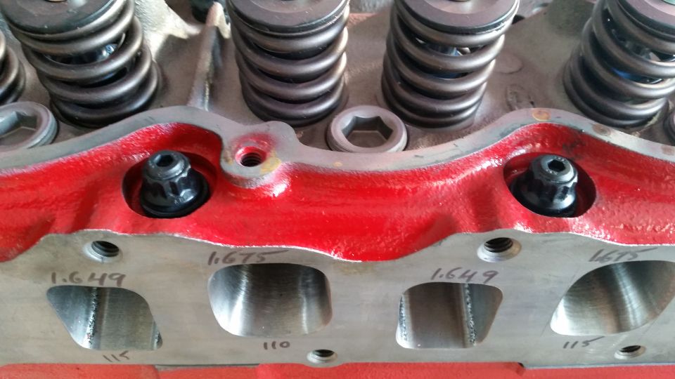 Close-up of a red engine cylinder head with black fasteners and size markings.