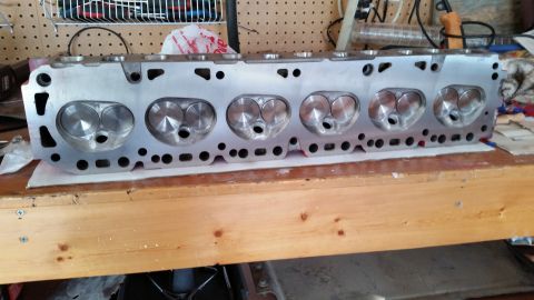 1981 Ford Bronco - A bare aluminum V8 engine cylinder head with six pairs of intake and exhaust valves is displayed on a wooden workbench. The cylinder head i…