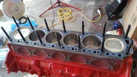1981 Ford Bronco - A red engine block is shown with a gasket and cylinder head studs installed. The gasket has blue sealing rings around the cylinder openings…