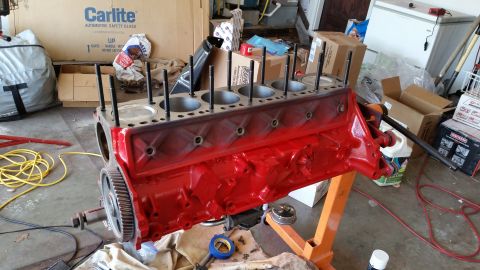 1981 Ford Bronco - A red V8 engine block is mounted on an orange engine stand, with cylinder head studs installed. Various car parts and boxes are visible in…