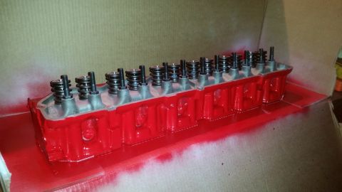 1981 Ford Bronco - A red-painted engine cylinder head with visible valve springs and bolts is shown from a 3/4 front angle. The part is resting on a cardboard…