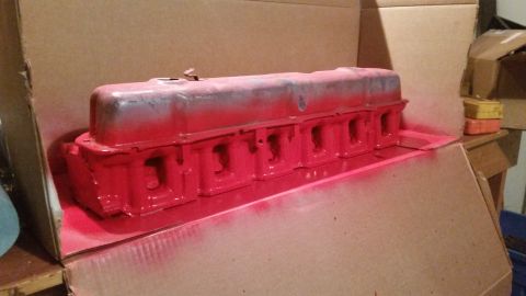1981 Ford Bronco - A red-painted engine valve cover is being spray-painted inside a cardboard box. The valve cover has a metallic gray primer visible in some…