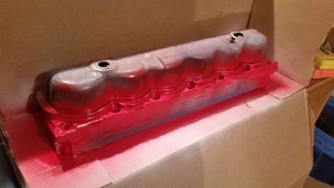 1981 Ford Bronco - A metal engine part, likely a valve cover, is being spray painted bright red inside a cardboard box. The part has a silver base coat and is…