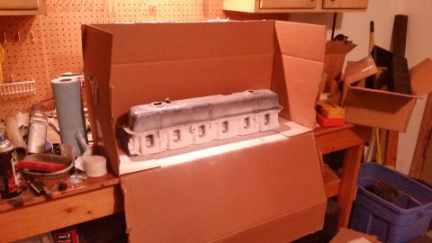 1981 Ford Bronco - A disassembled engine part, likely a valve cover, is placed inside a cardboard box. The part is silver and white and appears to be resting…