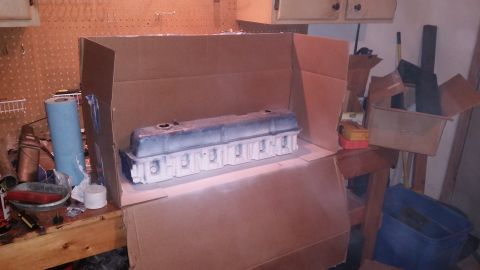 1981 Ford Bronco - A car engine valve cover is being spray painted inside a cardboard box setup in a garage. The valve cover is partially painted white and bl…
