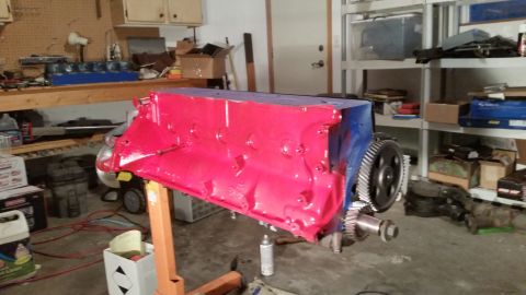 1981 Ford Bronco - A partially assembled engine block, painted bright red and blue, is mounted on an orange engine stand in a garage. Various tools and parts…