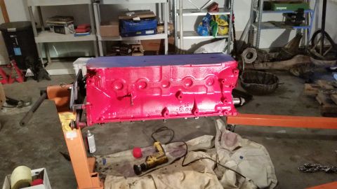 1981 Ford Bronco - A bright red engine block is mounted on an orange engine stand, with a blue tarp covering the top. Various tools and automotive parts are s…