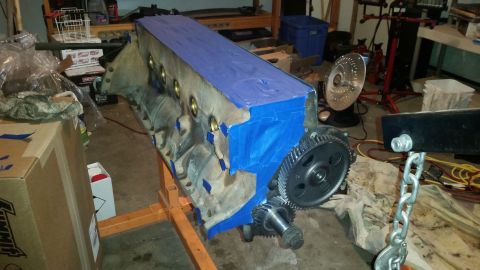 1981 Ford Bronco - A large engine block, partially covered in blue masking tape, sits on an orange engine stand. Visible gears are attached to the front of th…