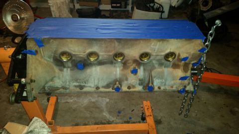 1981 Ford Bronco - A V8 engine block is mounted on an orange engine stand and covered with blue painter's tape. Several brass plugs are visible in the top of…