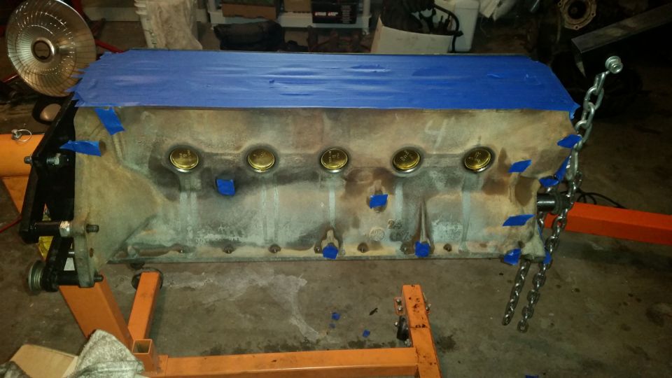 V8 engine block on an orange stand, covered in blue tape with brass plugs visible.