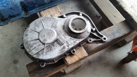 1981 Ford Bronco - A silver-colored metal automotive part, likely a timing cover, is resting on wooden planks. The part has a circular opening and embossed te…