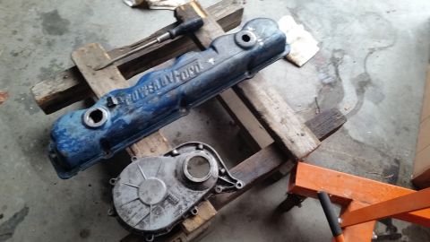 1981 Ford Bronco - A blue Ford valve cover and a silver timing cover are resting on wooden blocks on a concrete floor. The valve cover has raised lettering th…