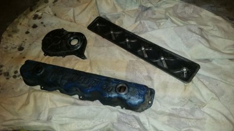 1981 Ford Bronco - Three automotive engine parts are laid out on a stained white cloth. Visible are a blue valve cover, a black timing cover, and a black gask…
