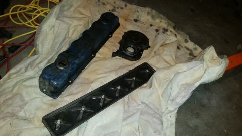 1981 Ford Bronco - A blue valve cover and two black engine parts are laid out on a stained white cloth on a concrete floor. Yellow and red electrical cords ar…