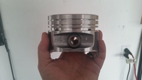 1981 Ford Bronco - A hand holds a single piston from a vehicle. The piston is silver and has several grooves for piston rings, and a hole for the wrist pin.