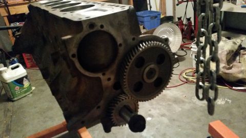 1981 Ford Bronco - A close-up view of an engine block with two gears attached, resting on an orange engine stand. A large metal chain hangs in the foreground.