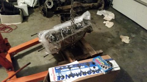 1981 Ford Bronco - Engine assembly covered in plastic, resting on a wooden pallet and an orange engine stand, with automotive gaskets in the foreground.