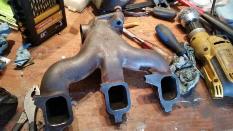 1981 Ford Bronco - A cast iron exhaust manifold sits on a workbench surrounded by tools, including a drill and pliers.