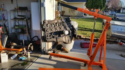 1981 Ford Bronco - An engine is suspended by a chain from an engine hoist, with a garage and residential area in the background.