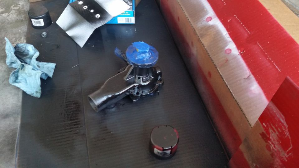 Car water pump with blue tape on top, sitting on a black surface next to a rag and some cans
