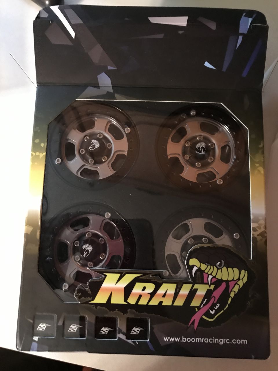 Four RC car wheels with silver rims and black tires in a box with a snake logo