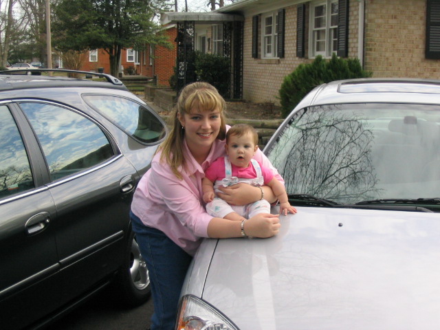 Woman holding a baby on the hood of a silver car, with a dark SUV parked next to it