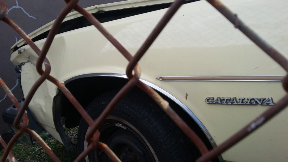 Light yellow car with 'CATALINA' badge behind a rusty chain-link fence, showing rear quarter panel damage.