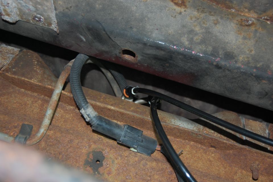 Undercarriage view of rusted metal frame with black electrical connector and wiring harness.