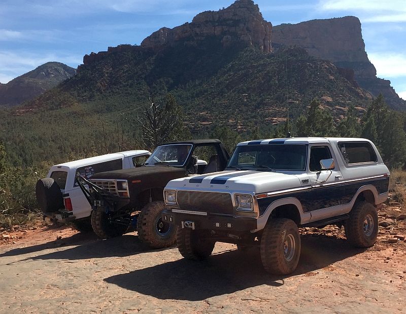 Three Ford Broncos parked on a dirt road with mountains in the background. The front Bronco is white with blue stripes.