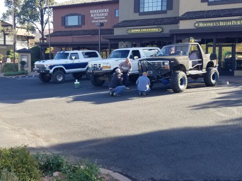 1995 Ford Bronco - Two lifted Ford Bronco SUVs are parked in front of a building with businesses. One is white with blue stripes and the other is dark gray, w…