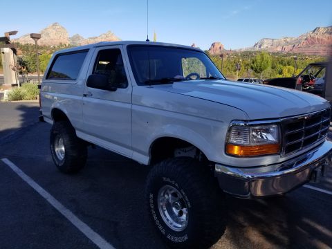 1995 Ford Bronco - A lifted white 1995 Ford Bronco is parked in a lot with red rock mountains in the background. The vehicle has large off-road tires and chro…