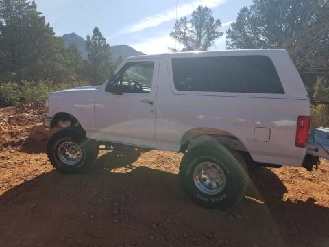 1995 Ford Bronco - A white 1995 Ford Bronco is parked on a dirt road with mountains and trees in the background. The vehicle has large off-road tires and appe…