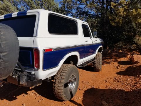 1995 Ford Bronco - The rear driver's side of a white and blue Ford Bronco is shown parked on a dirt road with trees in the background. The vehicle has a spare…